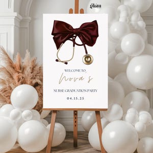 Puede incluir: Un letrero blanco sobre un caballete con un lazo burdeos y una ilustración de estetoscopio dorado. El letrero dice "WELCOME TO, Nora S NURSE GRADUATION PARTY 04.15.25". Globos blancos y hierba de la pampa en el fondo.