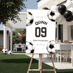 Puede incluir: Un letrero blanco sobre un caballete de madera con gráficos de balones de fútbol. El letrero dice "Bienvenido a Quinn 09" y "9º Cumpleaños de Quinn 05.18.25". Globos blancos y negros y balones de fútbol en el fondo.