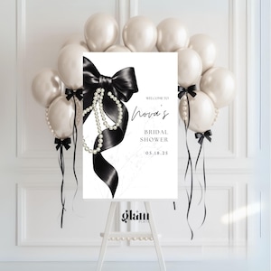 Puede incluir: Un letrero de bienvenida para una despedida de soltera con un lazo negro y detalles de perlas. El letrero dice "Welcome to Nova's Bridal Shower 05.18.25" y está rodeado de globos blancos y cintas negras. El letrero está sobre un caballete blanco.