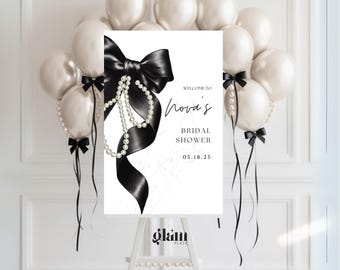 May include: A bridal shower welcome sign with a black bow and pearl accents. The sign reads "Welcome to Nova's Bridal Shower 05.18.25" and is surrounded by white balloons and black ribbons. The sign is on a white easel.