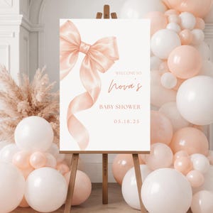 Puede incluir: Un letrero de bienvenida para baby shower sobre un caballete de madera. El letrero presenta un lazo y una cinta de acuarela en color melocotón. El texto en el letrero dice "Welcome to Nova's Baby Shower" y la fecha "05.18.25". Globos blancos y melocotón decoran el fondo.