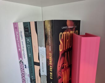 May include: A pink bookend supporting a stack of books. The bookend is made from a smooth, solid material. The books are of varying sizes and colours. The top book has the title "Strong and Wild" and the author "Sloane St. James".