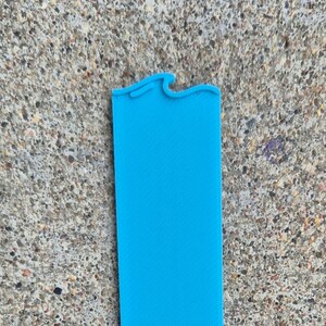 May include: A bright turquoise silicone watch band, rectangular in shape, with a wavy design at one end. The band is lying on a textured gray surface.