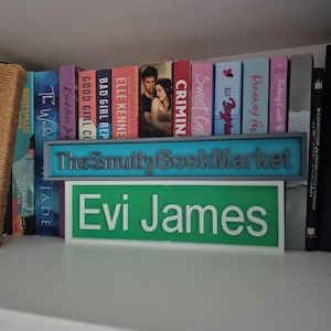 Op de afbeelding: Een boekenplank toont een verzameling boeken met kleurrijke ruggen. Een bord met de tekst "The SmuttyBookMarket" is zichtbaar, samen met een groen bord met "Evi James". De boeken zijn gerangschikt op een witte plank.