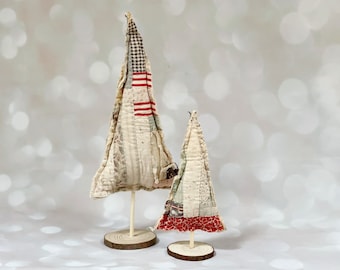 May include: Two fabric Christmas trees on wooden bases. The larger tree is white with red and blue patchwork fabric. The smaller tree is white with red and green patchwork fabric.