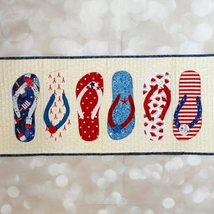 May include: A quilted table runner featuring six flip-flop designs in red, white, and blue patterns. The flip-flops have various patriotic motifs, including stars, stripes, and fireworks, on a cream background with a navy blue border.