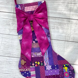 May include: A purple and pink patchwork Christmas stocking with a large pink bow at the top. The stocking is made of various fabrics with different patterns and textures. The word "MOSCATO" is visible on the stocking.