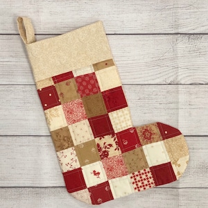May include: A red and cream patchwork Christmas stocking with a cream fabric cuff. The stocking is made of squares of different fabrics with floral and geometric patterns.