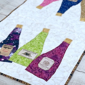 May include: A quilted table runner with a white background and a border of brown and black stripes. Five wine bottles are arranged vertically on the runner, each with a different colored fabric and a vintage-style label. The labels include the text "Moulin a Vent", "Chateau de Cossiat", "Moulin Detesson", and "Le Royer de la Pouget".