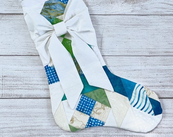 May include: A white Christmas stocking with a large white bow. The stocking is made of patchwork fabric in shades of blue, green, and white.