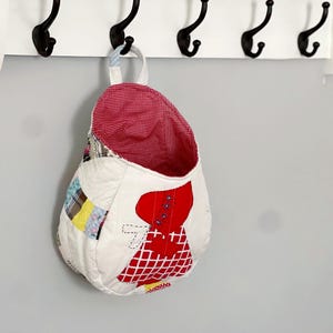 May include: A white fabric hanging storage bag with a red and white checkered lining. The bag is decorated with a patchwork design and a red applique of a woman in a dress.