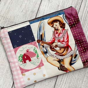 May include: A patchwork fabric zippered pouch with a colorful design featuring a woman playing a guitar, a horseshoe, a barn, and polka dots.