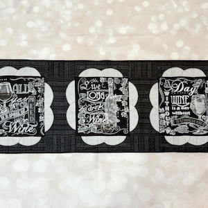 May include: A black and white wine-themed table runner with three rectangular panels. Each panel features a different wine-related quote in elegant script, with images of wine glasses and bottles. The runner is bordered with a decorative pattern.