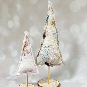 May include: Two white fabric Christmas trees on wooden bases. The larger tree is decorated with a patchwork of colorful fabrics. The smaller tree is plain white.
