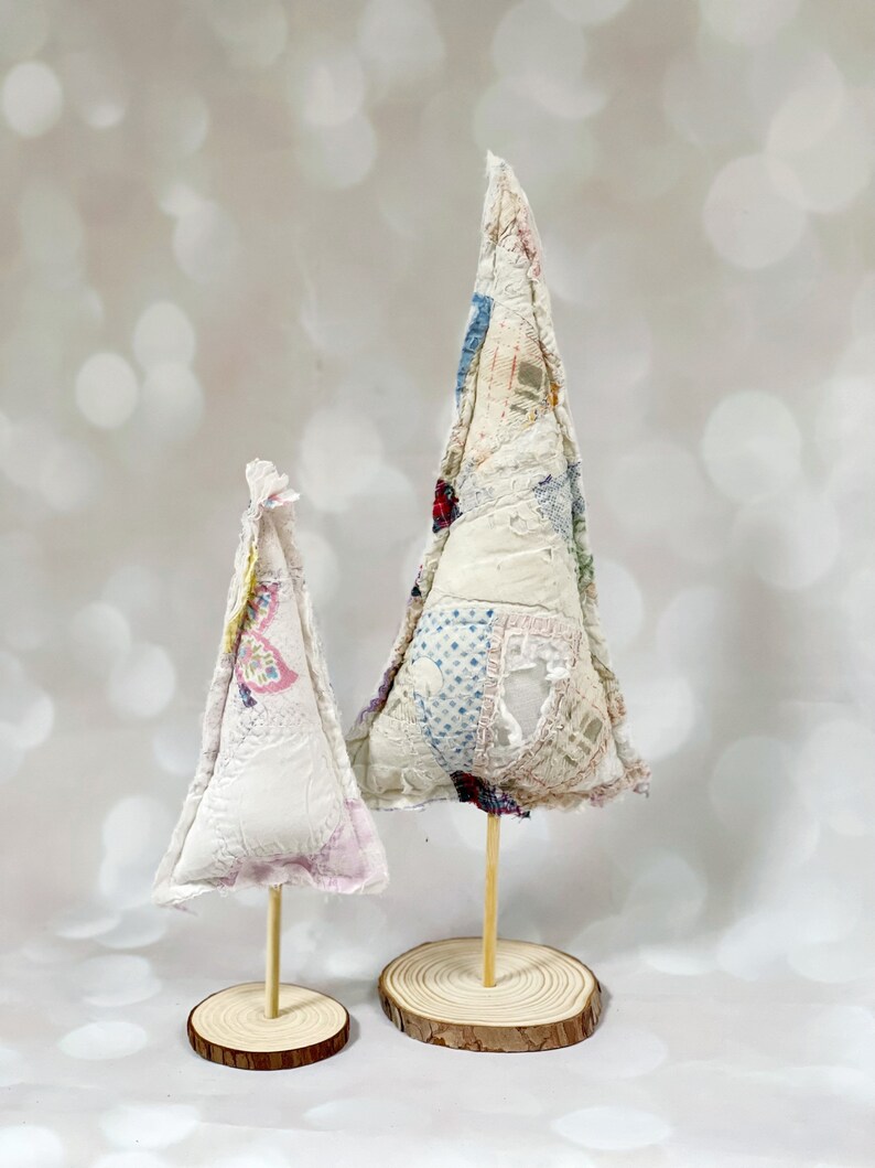 May include: Two white fabric Christmas trees on wooden bases. The larger tree is decorated with a patchwork of colorful fabrics. The smaller tree is plain white.
