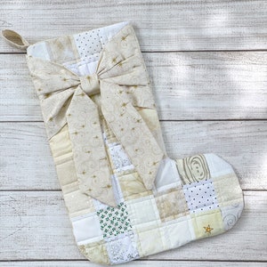 May include: A white and cream patchwork Christmas stocking with a gold bow and gold star accents.
