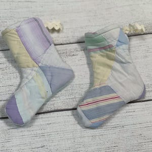 May include: Two handmade patchwork Christmas stockings in a light blue, purple, and yellow color scheme. The stockings are made from repurposed fabric and have a unique, rustic look.