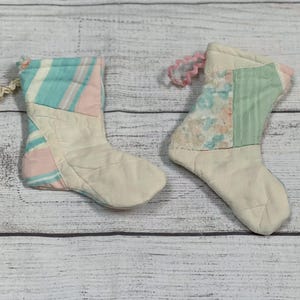 May include: Two patchwork fabric Christmas stockings in pink, green, and blue. The stockings are hanging on a white wooden background.