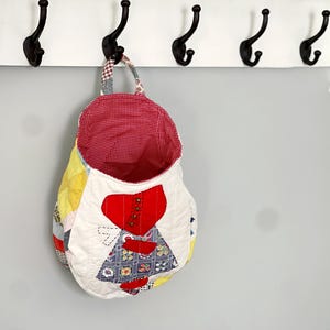 May include: A patchwork fabric hanging storage bag with a red heart design and red and white gingham lining. The bag is hanging on a white shelf with black hooks.