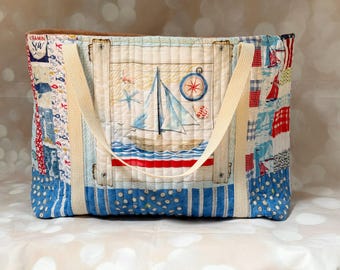 May include: A quilted tote bag featuring a nautical theme with a central sailboat illustration, a compass, and a starfish. The fabric displays a patchwork design with blue, red, and white patterns, including stripes and text that reads Vitamin Sea.