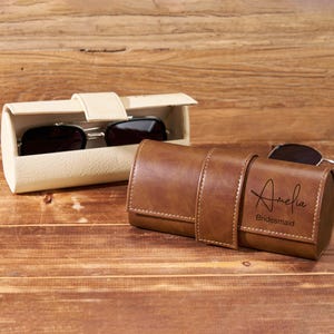 May include: A brown leather sunglasses case with the name "Amelia" and the word "Bridesmaid" engraved on the front. A pair of sunglasses rests on the wooden surface next to the case. Another off-white sunglasses case is open, revealing a pair of sunglasses.