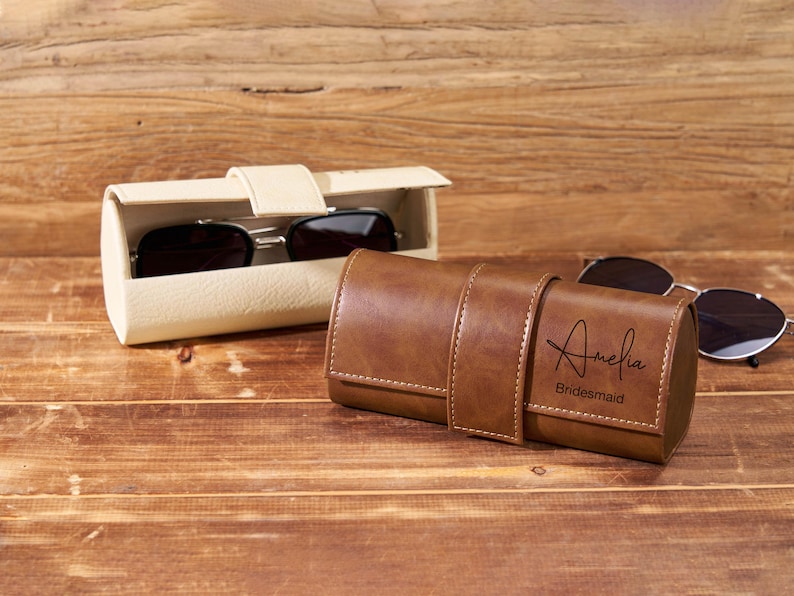 May include: A brown leather sunglasses case with the name "Amelia" and the word "Bridesmaid" engraved on the front. A pair of sunglasses rests on the wooden surface next to the case. Another off-white sunglasses case is open, revealing a pair of sunglasses.