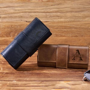 May include: Two leather eyeglass cases, one navy blue and one brown, rest on a wooden surface. The brown case is personalized with the name "Albert". Sunglasses are placed on the right side of the image.
