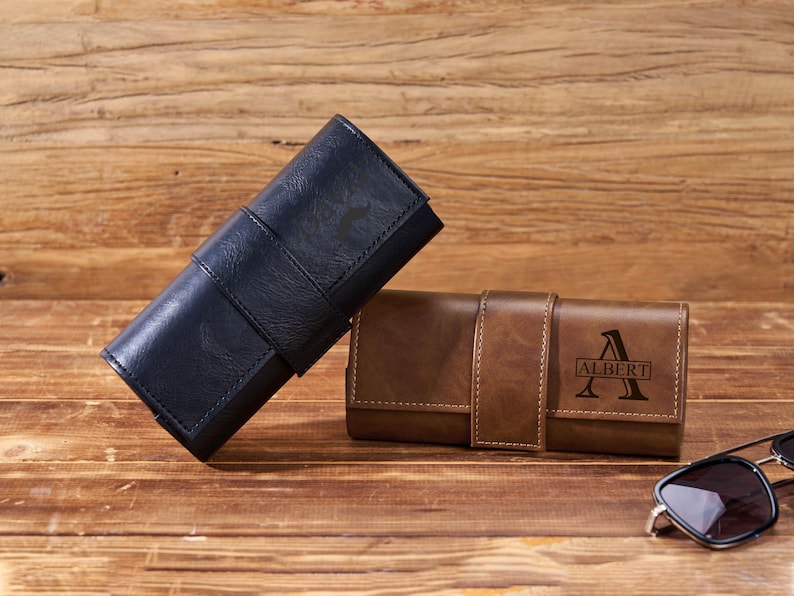 May include: Two leather eyeglass cases, one navy blue and one brown, rest on a wooden surface. The brown case is personalized with the name "Albert". Sunglasses are placed on the right side of the image.