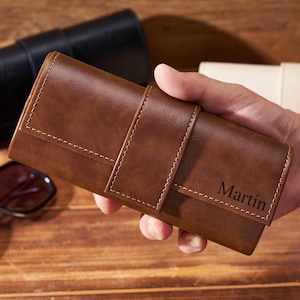 May include: A brown leather watch roll with white stitching and a strap closure is held in a hand. The word "Martin" is printed on the front. Sunglasses and a black watch case are in the background.
