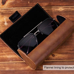 May include: Black sunglasses with a silver frame rest inside a brown leather case with a black interior. The case is open, revealing the glasses and a black, rectangular storage area. The text "Flannel lining to protect glasses" is visible.