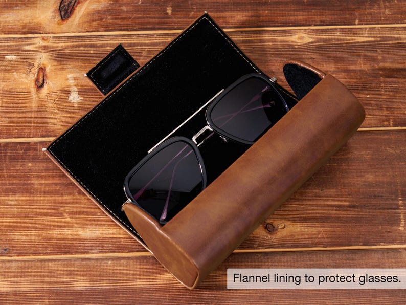 May include: Black sunglasses with a silver frame rest inside a brown leather case with a black interior. The case is open, revealing the glasses and a black, rectangular storage area. The text "Flannel lining to protect glasses" is visible.