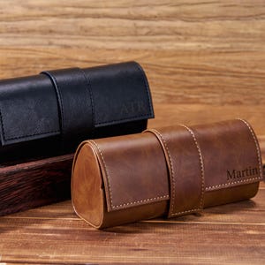 May include: Two leather eyeglass cases, one black and one brown, sit on a wooden surface. The black case has "ATR" embossed on the flap, while the brown case has "Martin" embossed. Sunglasses are partially visible.