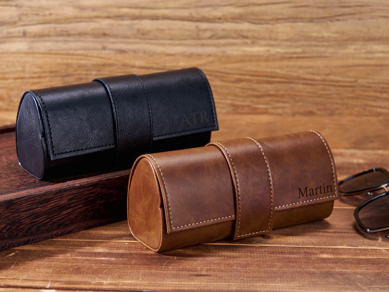 May include: Two leather eyeglass cases, one black and one brown, sit on a wooden surface. The black case has "ATR" embossed on the flap, while the brown case has "Martin" embossed. Sunglasses are partially visible.