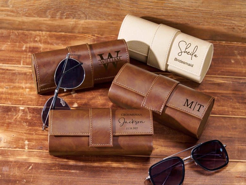 May include: Several personalized brown leather sunglasses cases with sunglasses. One case is cream-colored. The cases are engraved with names, initials, and the word "Groomsman" with a date. The sunglasses have dark lenses and silver frames.