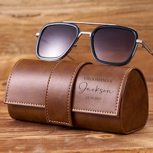 May include: Sunglasses with a black frame and gradient lenses rest on a brown leather case. The case is embossed with the word "Groomsman", a name, and a date. The case is secured with a matching leather strap.