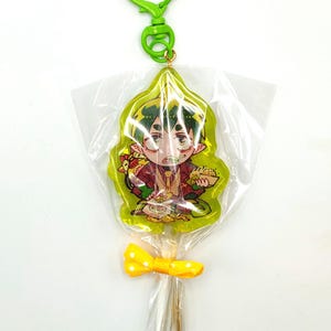 May include: A green keychain with a cartoon character lollipop design. The lollipop is wrapped in clear cellophane and tied with a yellow bow. The keychain has a green heart-shaped clasp.