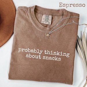 Pode incluir: Camiseta cor espresso com a frase "probably thinking about snacks" em letras brancas. A camiseta tem gola redonda e uma etiqueta pequena. Um colar de prata com um pingente está sobre a camiseta.