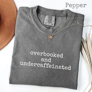 Könnte beinhalten: Ein graues Comfort Colors T-Shirt mit dem weißen Text "overbooked and undercaffeinated". Eine goldene Anhänger-Halskette liegt auf dem Shirt. Das Shirt ist gefaltet, und ein brauner Hut ist teilweise im Hintergrund sichtbar.