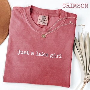 May include: Crimson-colored t-shirt with the text "just a lake girl" in white. The shirt has a crew neck and a small tag on the upper right. A silver necklace with a pendant and a brown hat are also visible.