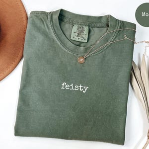 May include: A moss green t-shirt with the word "feisty" embroidered on the front. The shirt is made of a soft, comfortable fabric and has a relaxed fit.