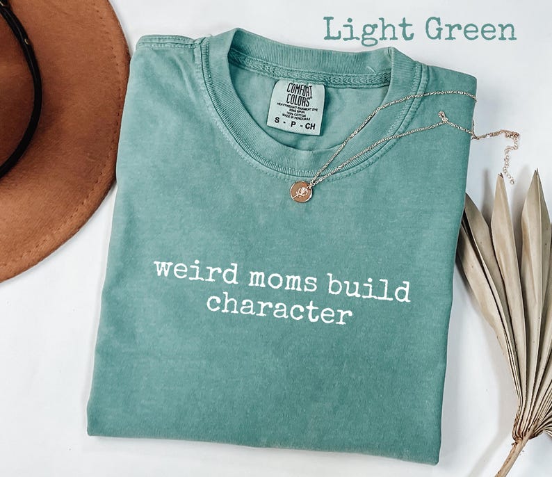 May include: Light green t-shirt with the text "weird moms build character" in white. A gold necklace with a pendant rests on the shirt. The shirt is folded, and a brown hat is partially visible in the background.