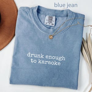 May include: A blue jean colored t-shirt with the phrase "drunk enough to karaoke" in white lettering. A gold necklace with a pendant is draped across the shirt. The shirt is folded on a white surface.