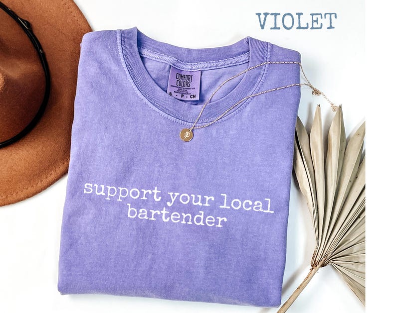 May include: A violet Comfort Colors t-shirt with the text "support your local bartender" in white. A gold necklace with a pendant is draped on the shirt. A brown hat is visible in the upper left corner.