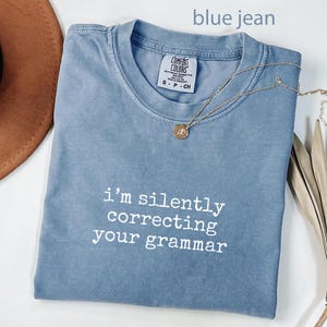 Peut inclure: Un t-shirt bleu jean avec le texte "I'm silently correcting your grammar" en blanc. Un collier en or avec un pendentif est drapé sur le t-shirt. Le t-shirt est plié sur une surface blanche.