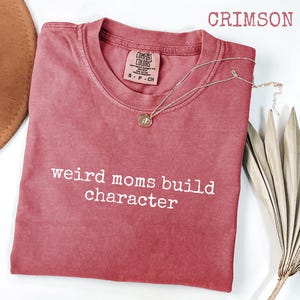 May include: Crimson-coloured t-shirt with the text "weird moms build character" in white. The shirt has a crew neck and a small tag on the upper right. A silver necklace with a pendant is draped across the shirt. A brown hat is partially visible.