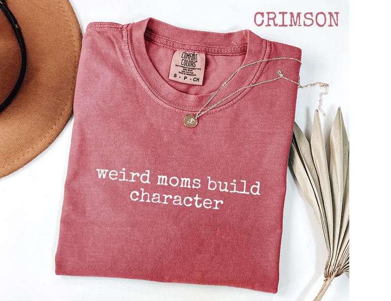 May include: Crimson-coloured t-shirt with the text "weird moms build character" in white. The shirt has a crew neck and a small tag on the upper right. A silver necklace with a pendant is draped across the shirt. A brown hat is partially visible.