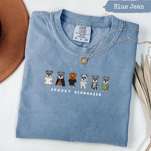 Puede incluir: Una camiseta color azul jean con un gráfico de seis perros Schnauzer con varios disfraces de Halloween, incluyendo un fantasma, vampiro, calabaza, momia, esqueleto y un suéter verde. El texto debajo de los perros dice SPOOKY SCHNAUZER.