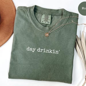 May include: A green t-shirt with the text "day drinkin'" in white lettering. The shirt is made of a soft, comfortable fabric and has a relaxed fit.