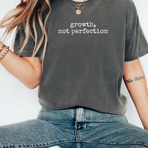 May include: Dark gray t-shirt with the text "growth, not perfection" in white. The shirt is a relaxed fit and is paired with blue jeans. A gold necklace and bracelet are also visible.