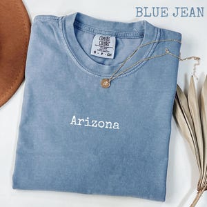 May include: A blue jean colored t-shirt with the word "Arizona" embroidered on the front. The shirt has a crew neck and short sleeves. A gold necklace with a pendant is draped across the shirt. The words "BLUE JEAN" are printed at the top.
